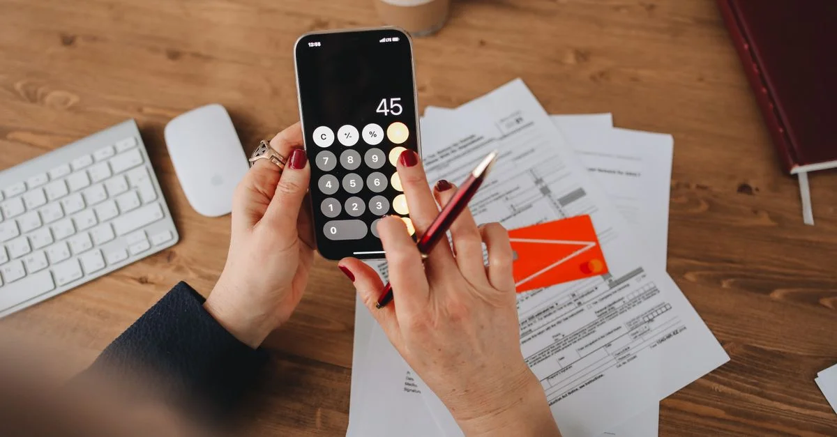 Des mains utilisant une application de calculatrice sur un smartphone au-dessus de documents financiers. Parfait pour illustrer des thèmes financiers ou technologiques.