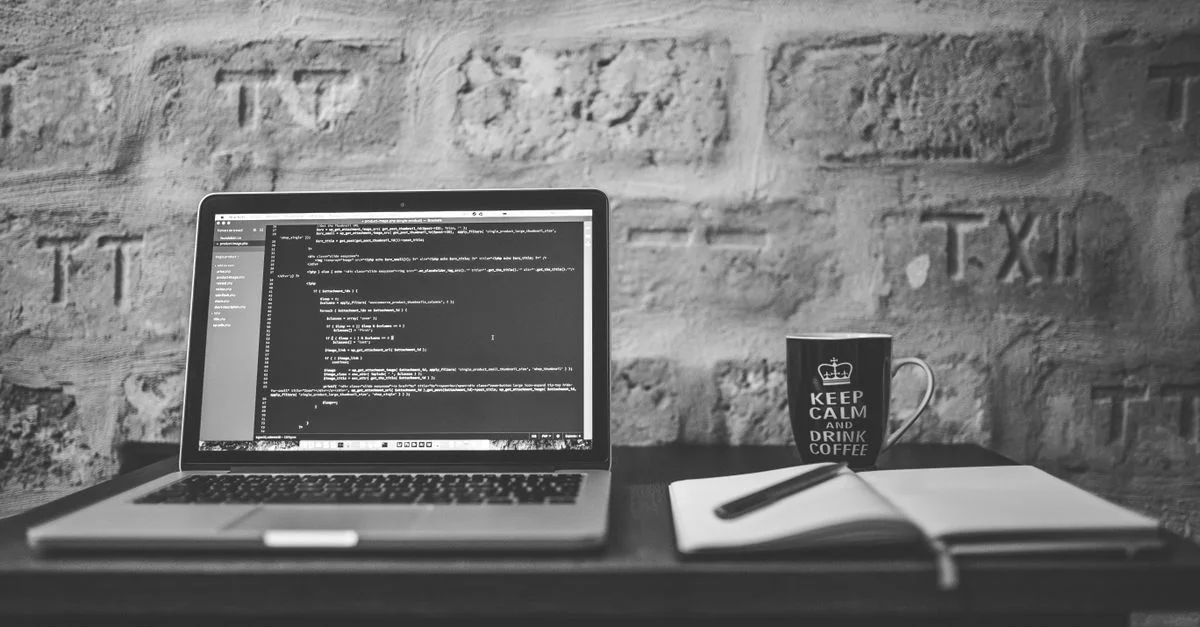 Photo en noir et blanc d'un espace de travail de codage avec un ordinateur portable, une tasse et un carnet.