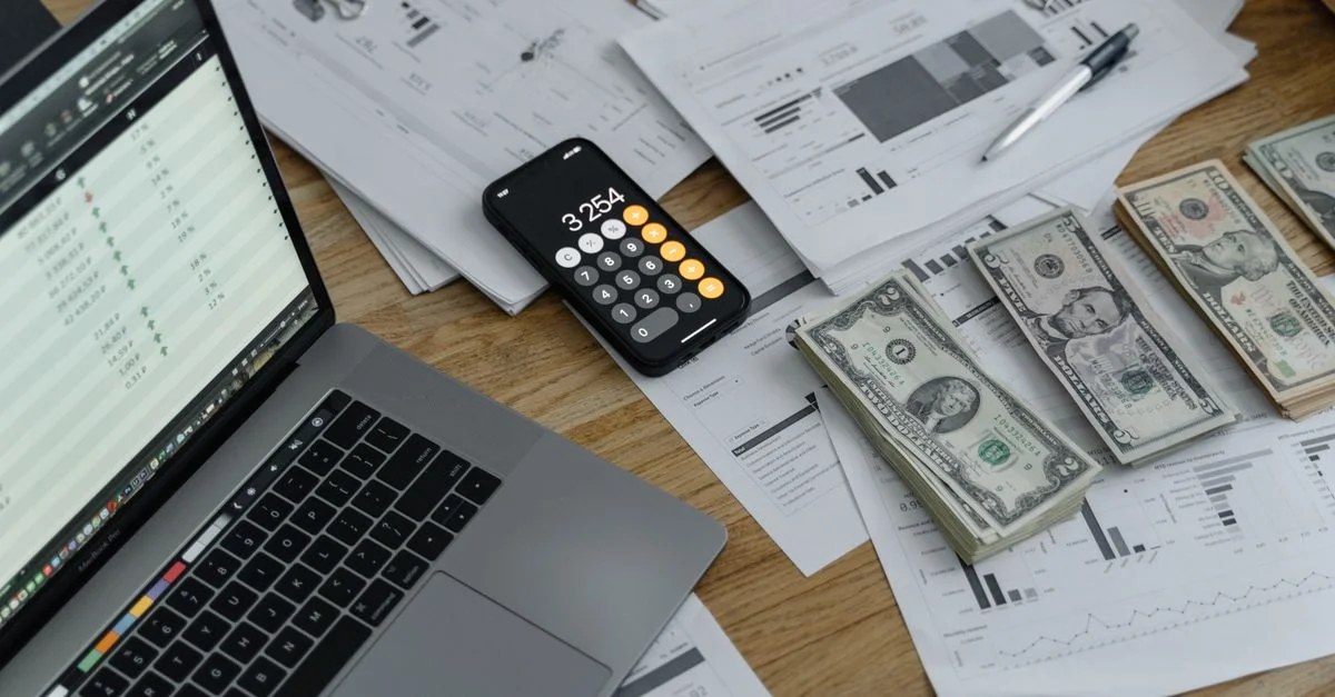 Un bureau avec des documents financiers, de la monnaie, un ordinateur portable et un calculateur de téléphone.
