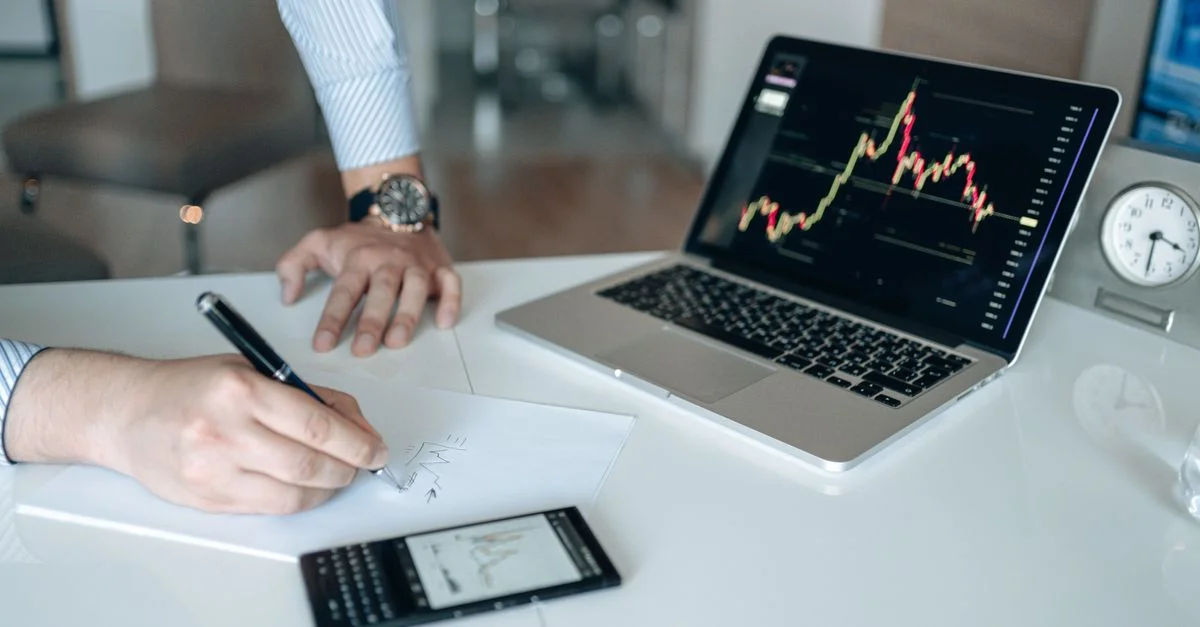 Espace de travail professionnel avec un ordinateur portable affichant des graphiques financiers et une personne qui écrit.