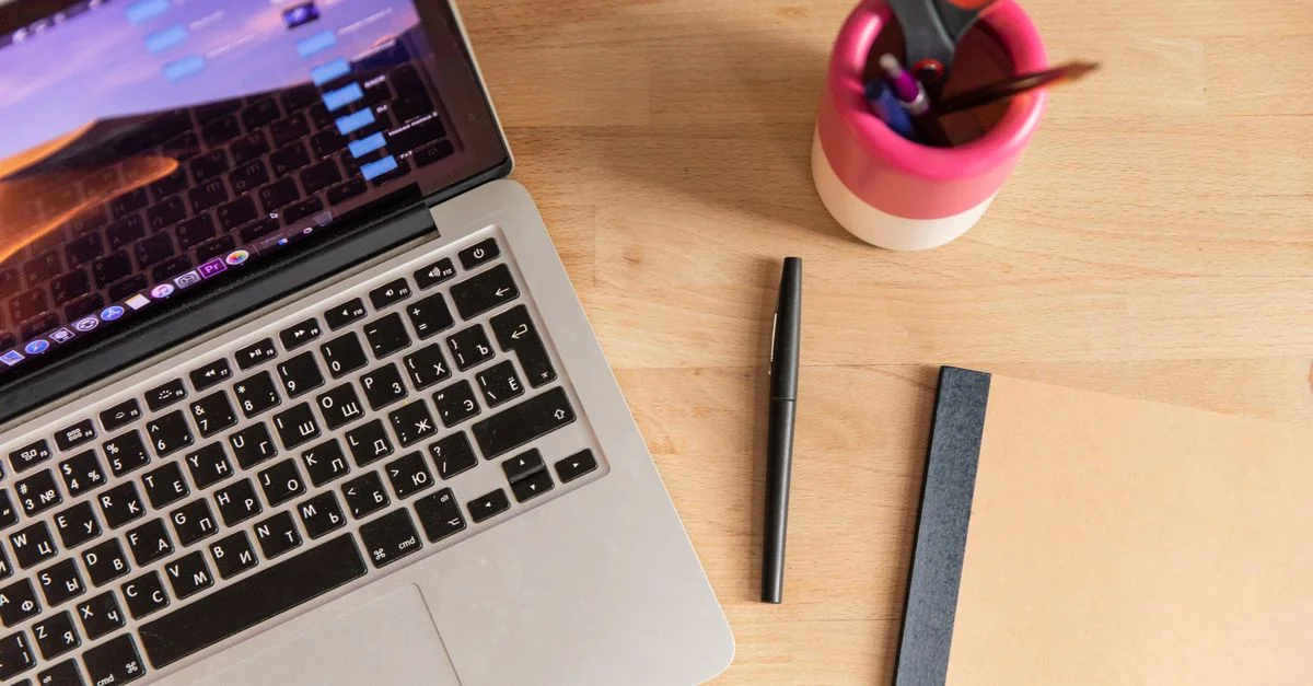 Vue de dessus d'un ordinateur portable, d'un stylo, d'un carnet et de ciseaux sur un bureau en bois, représentant un espace de travail.