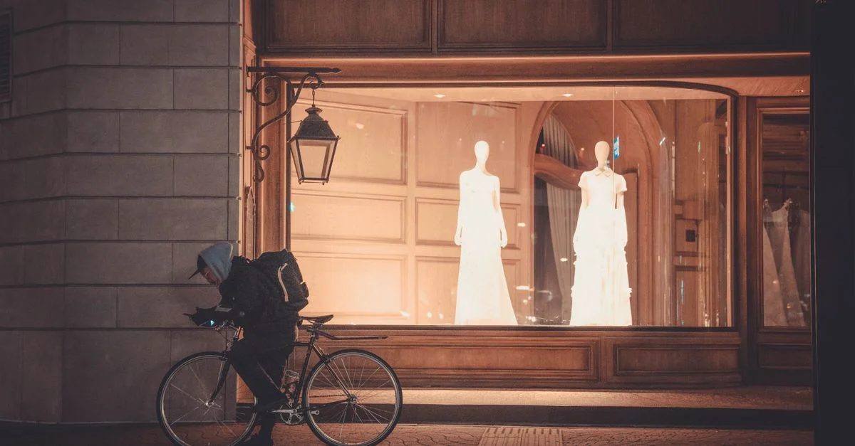 Un cycliste passe devant une vitrine éclairée montrant des mannequins en robes de mariée la nuit.
